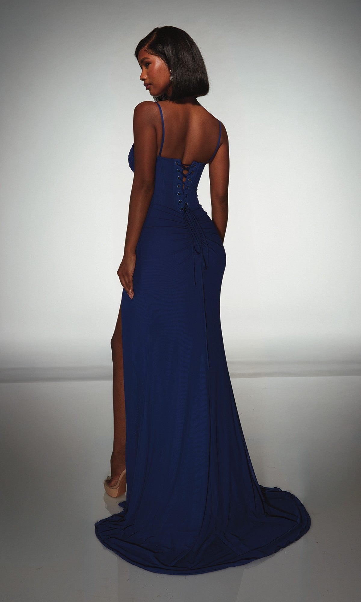 A woman faces away from the camera, standing on a reflective surface in the Alyce 62002 long prom dress, featuring a dark blue color, corset bodice, lace-up back, and thigh-high slit.